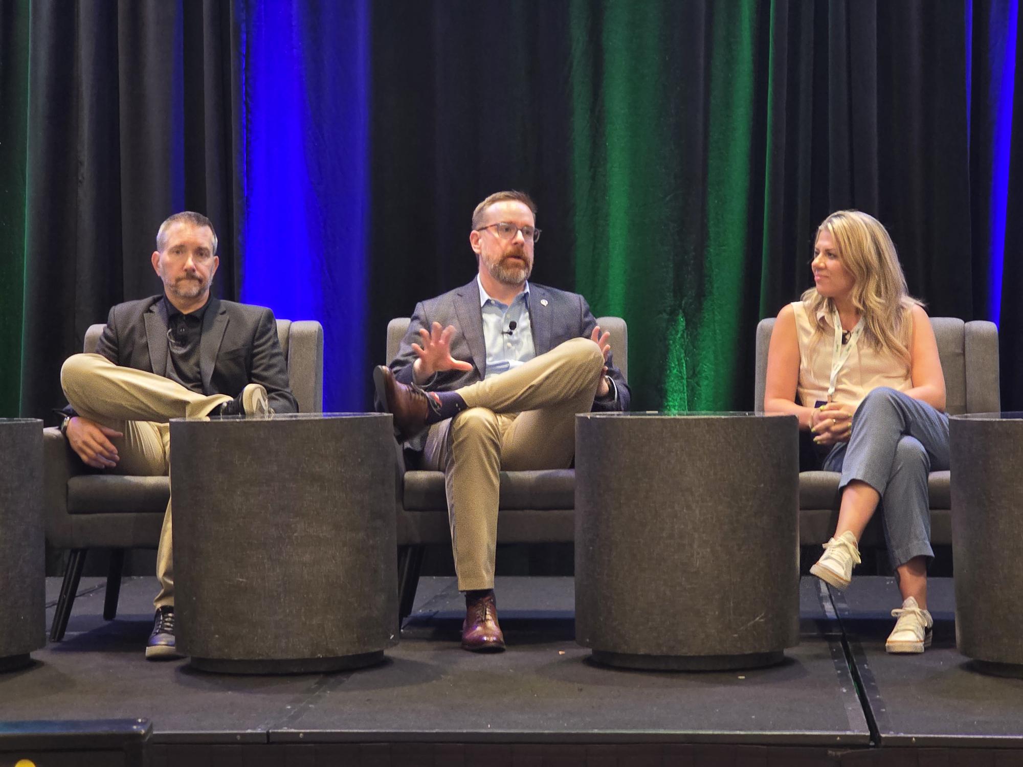 Three professionals participate in a panel discussion on stage with blue and green lighting backdrop