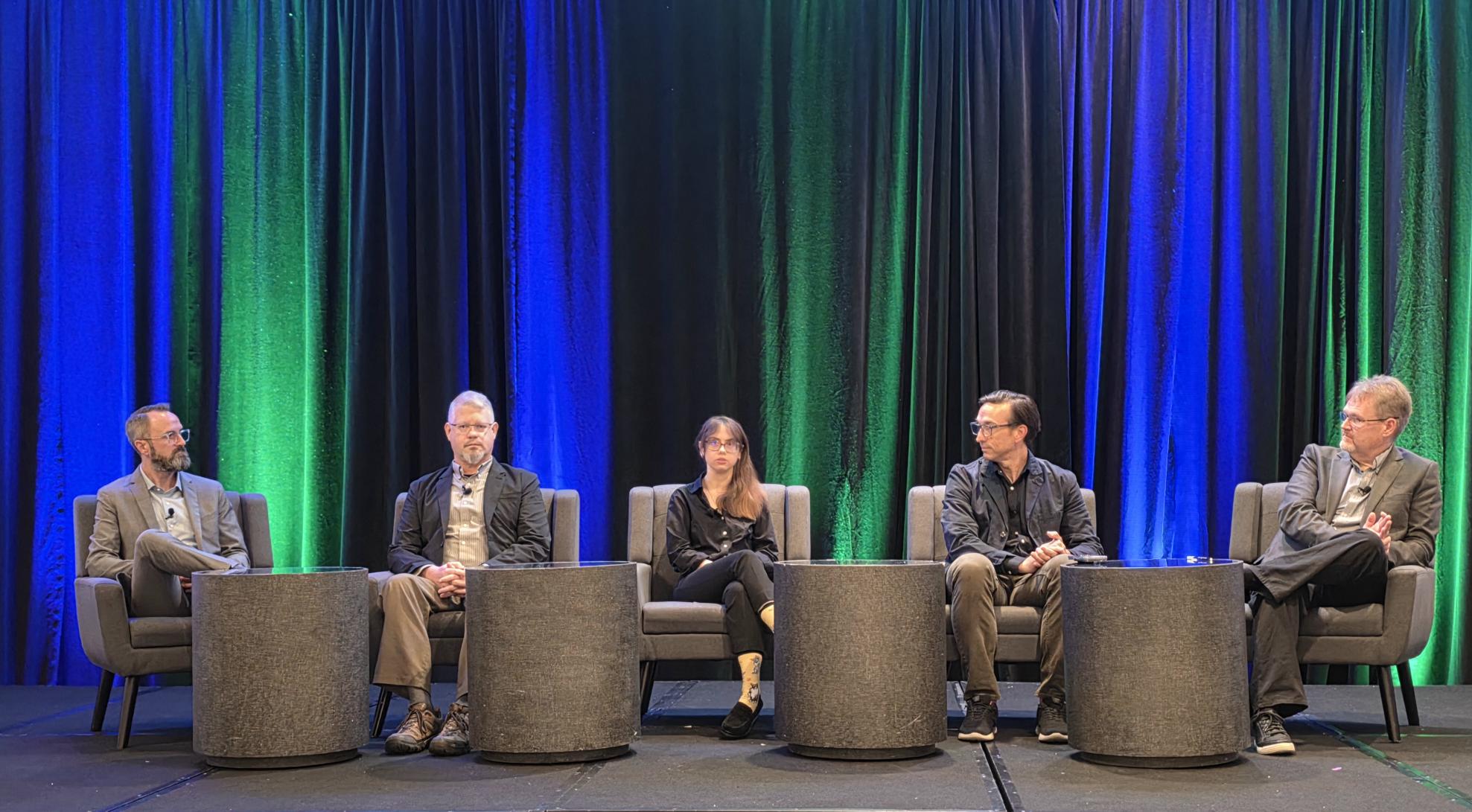 Five professionals seated on stage during a panel discussion with blue and green curtains in background
