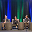 Five professionals seated on stage during a panel discussion with blue and green curtains in background