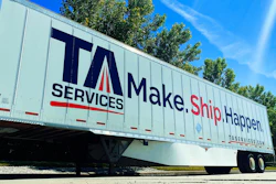 TA Services semi-trailer with 'Make.Ship.Happen' tagline parked against blue sky and trees