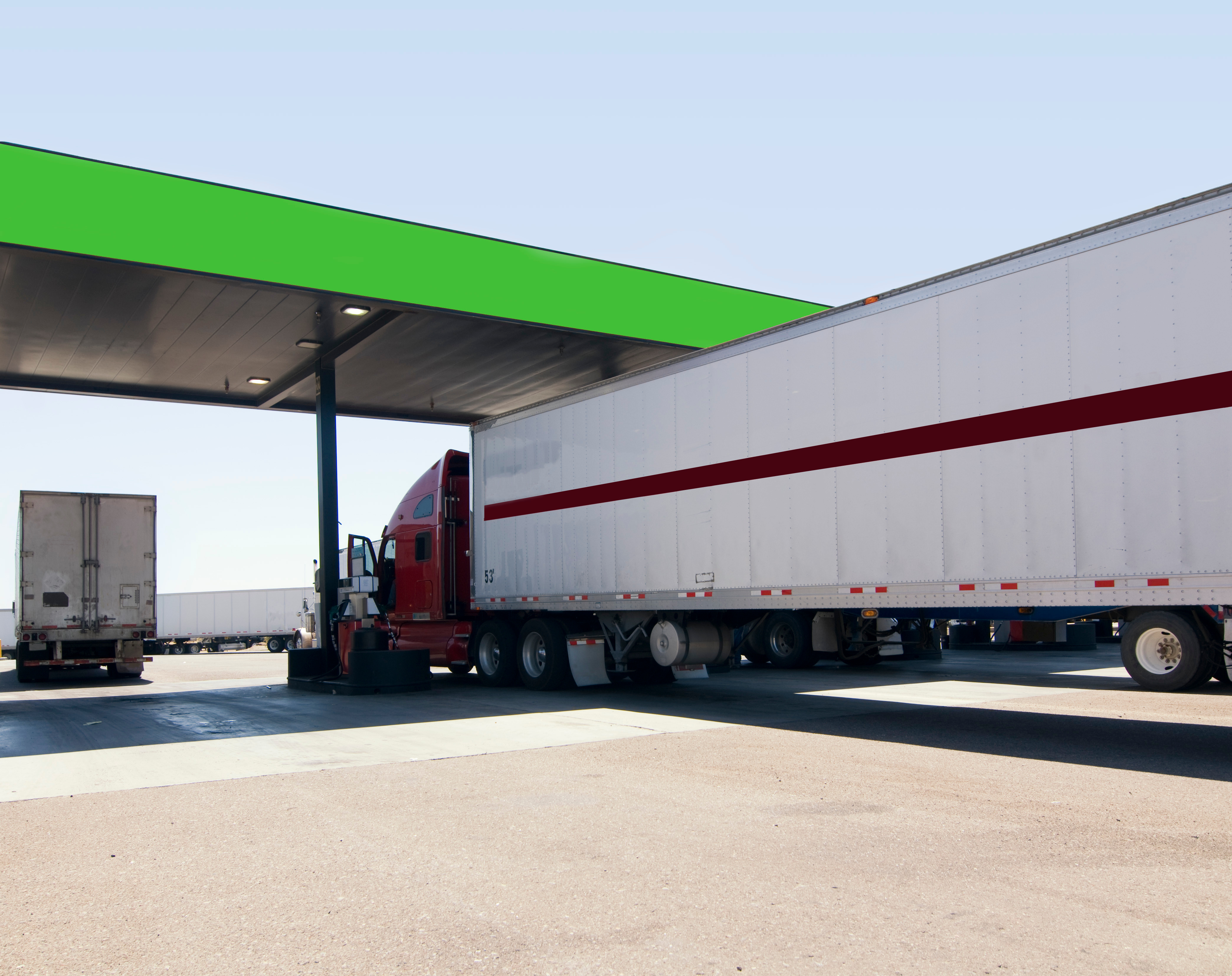 Red semi-truck with white trailer parked under green canopy at industrial loading dock facility