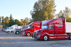 Fleet of red and white semi-trucks parked in a row at a truck stop with autumn trees in the background