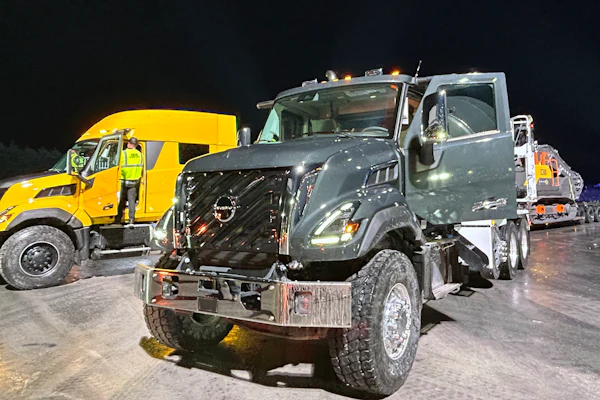 Commercial trucks lined up at night, including yellow Volvo and gray Nissan vehicles in fleet formation