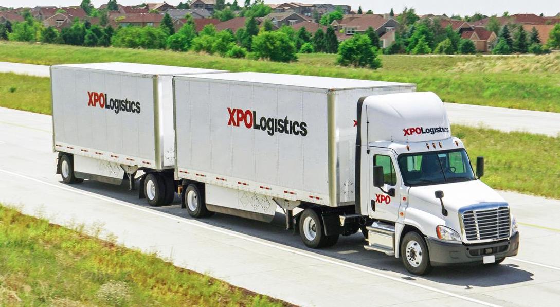 XPO Logistics semi-truck with double trailers driving on highway through suburban landscape